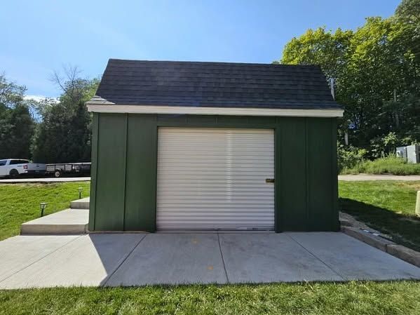 Small green shed with a white roll-up door on a concrete pad in a grassy yard.