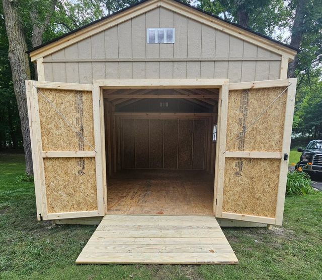 A wooden shed with open double doors and a ramp, set on grass. The shed is empty inside.