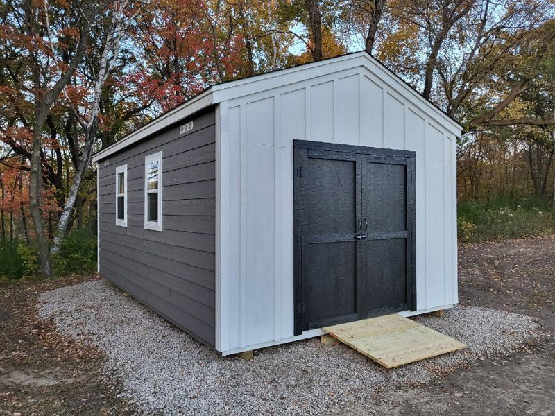 Small gray and white storage shed with double doors, sitting on gravel in a wooded yard.