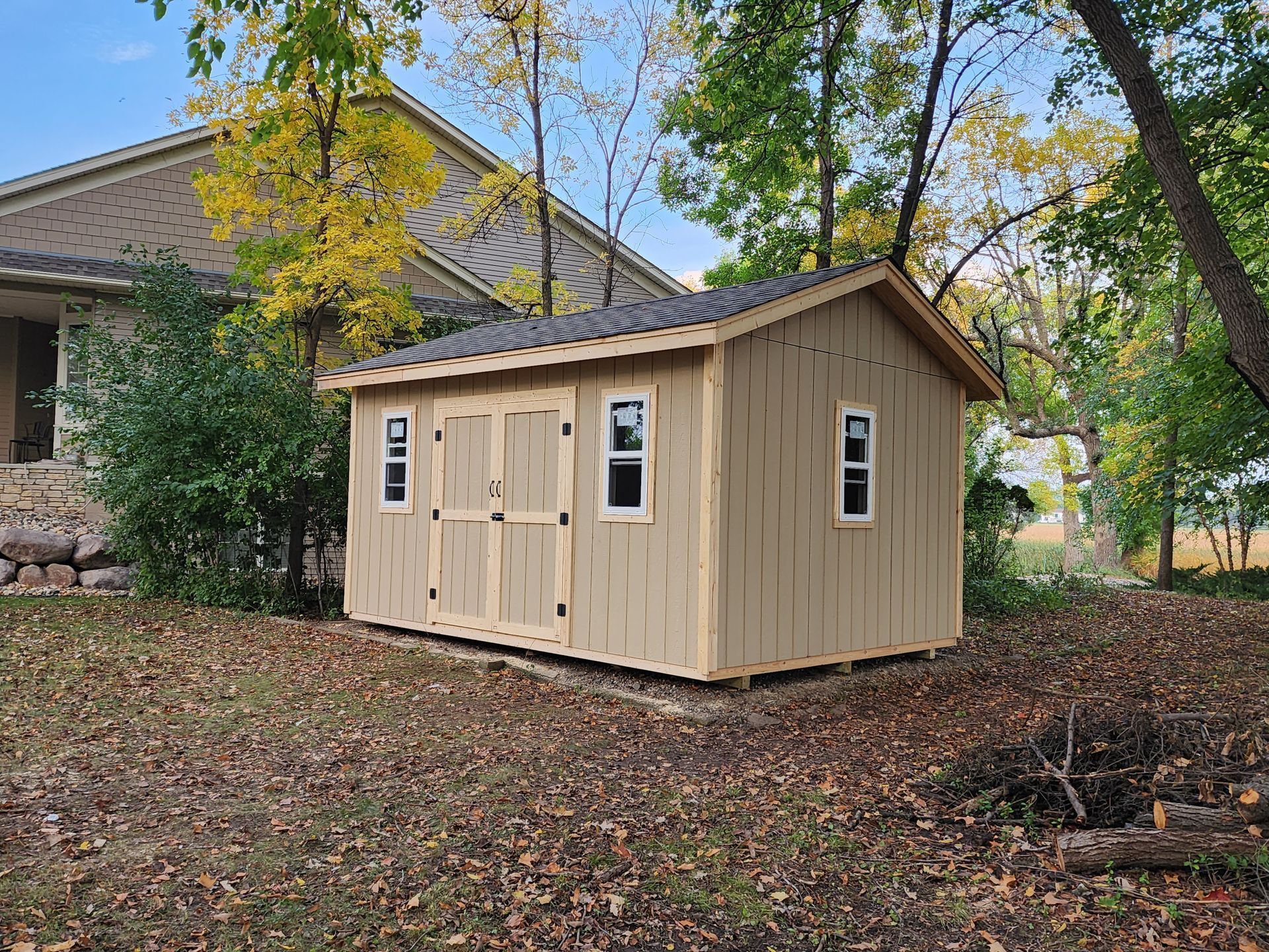 Small beige shed with dark roof in a leaf-covered yard beside a house and trees