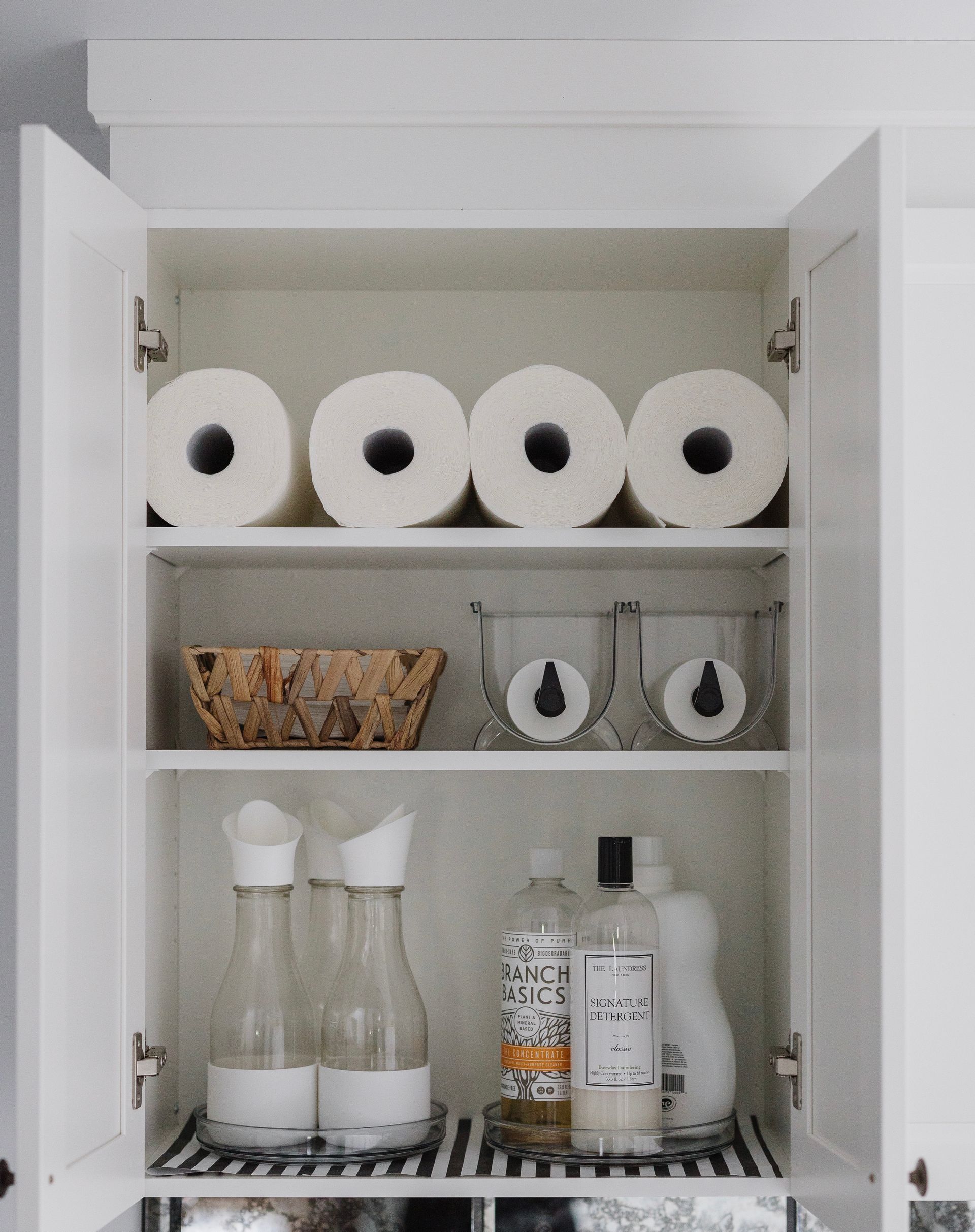 White cabinet interior with neatly arranged cleaning supplies, paper towels, and decorative jars.