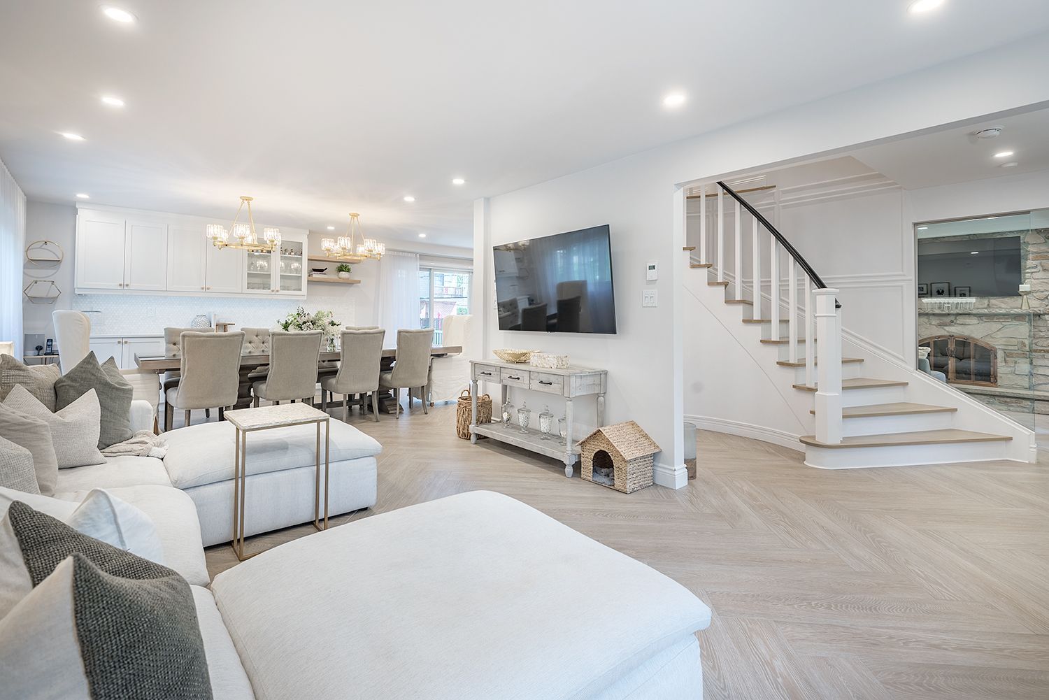 Open-concept living space with white walls, light wood floors, and a modern staircase. Dining area and sofa visible.