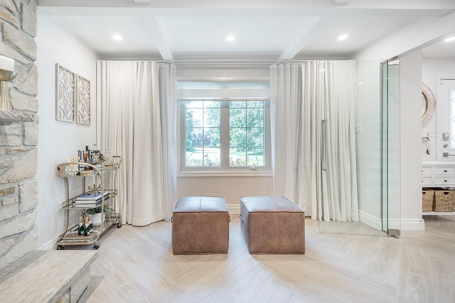 White-walled master bathroom with a window, two ottomans, a glass shower, and vanity.