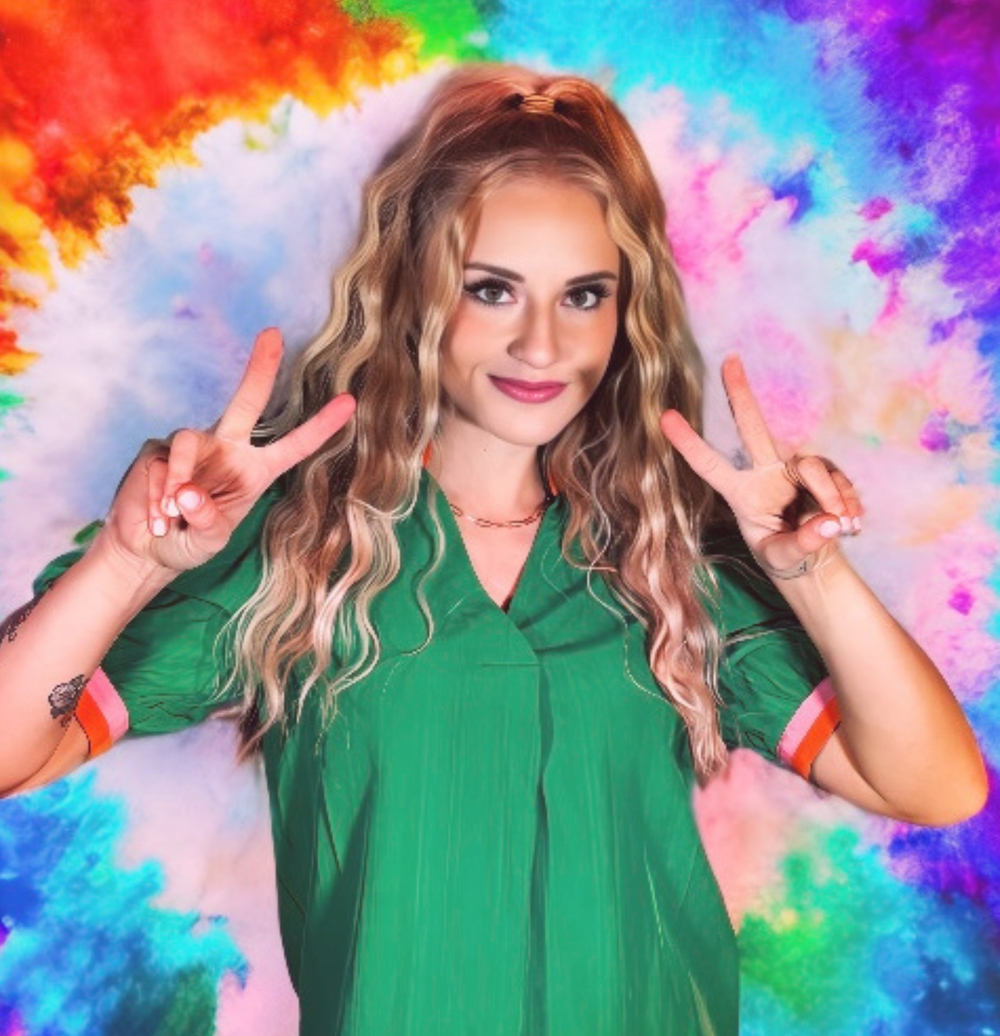 Woman with blonde wavy hair makes peace signs in front of a tie-dye background.