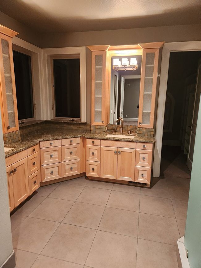 Empty bathroom vanity area with light wood cabinets, granite countertop, mirror, and beige tile floor.