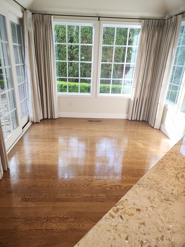 Empty sunlit room with hardwood floors, bay windows, and beige patterned curtains