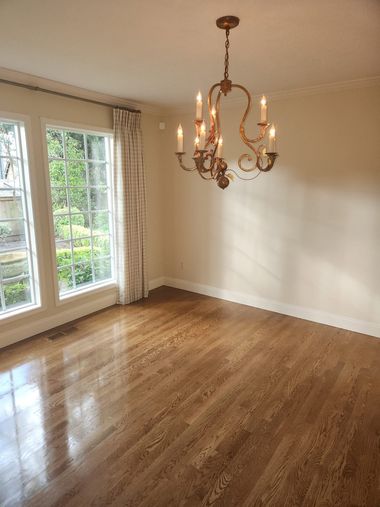 Empty beige room with hardwood floor, large windows, and a brass chandelier hanging from the ceiling