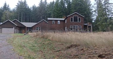 Rustic wooden house and garage beside a grassy field with tall evergreen trees in the background.