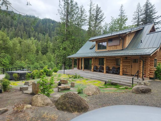 Rustic log cabin with green roof in a rocky forest clearing, surrounded by trees and mountains.