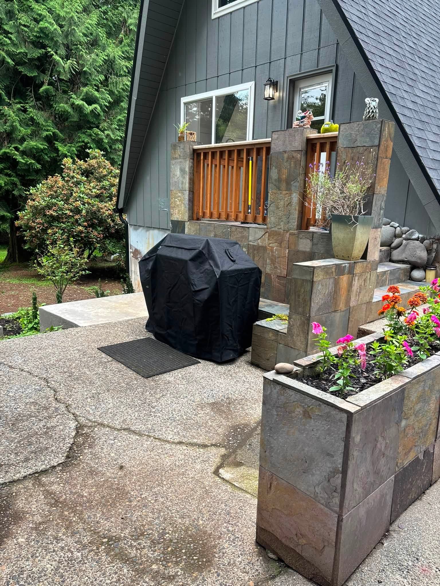 Small patio with a covered grill, flower planters, and a modern gray house wall with windows.