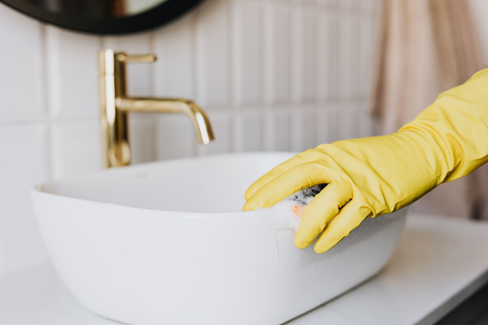 Person wearing yellow gloves cleaning a white bathroom sink with a sponge.
