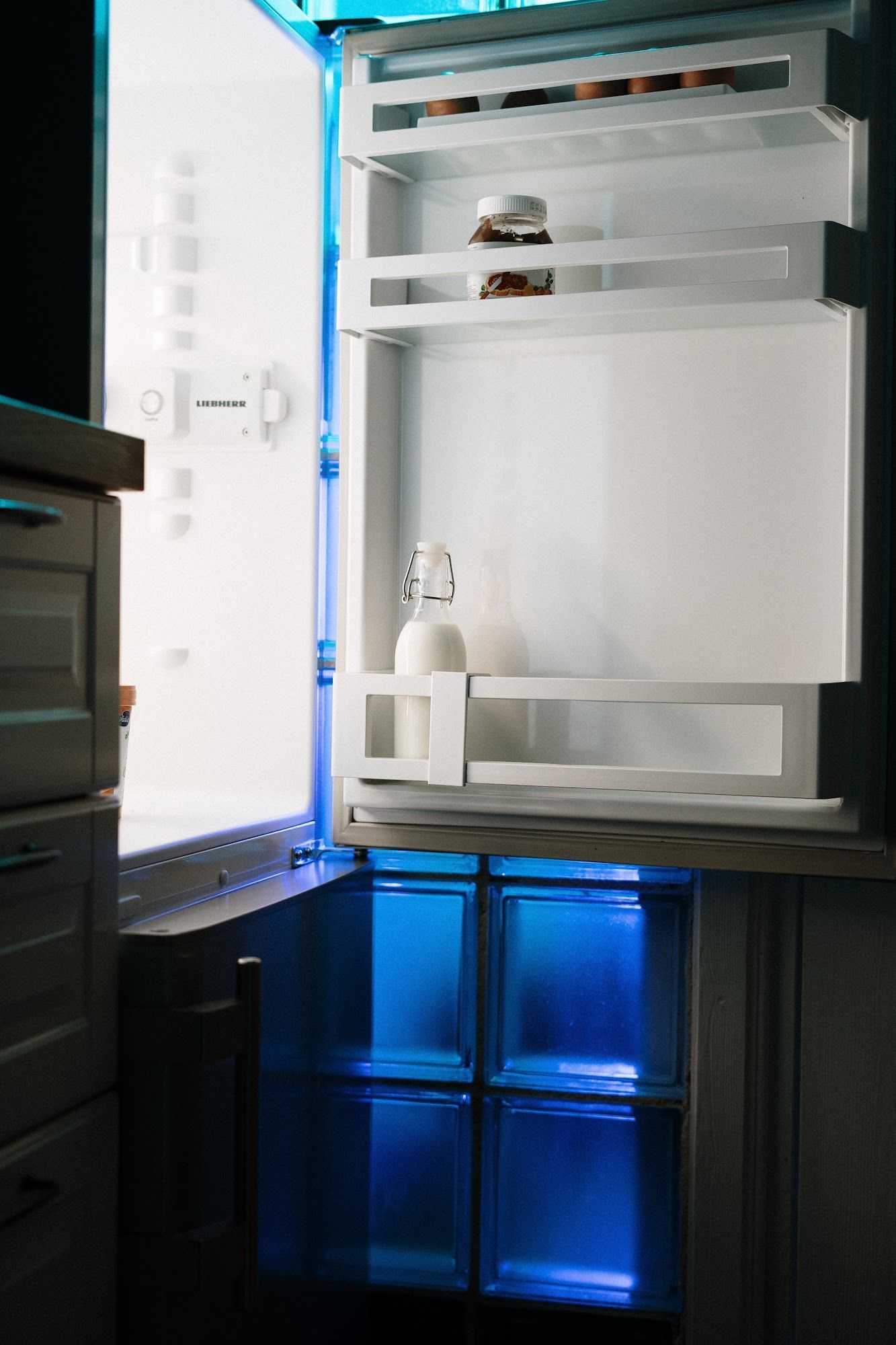 Refrigerator with the door open, illuminated with blue and white lights. Inside, a bottle is visible.