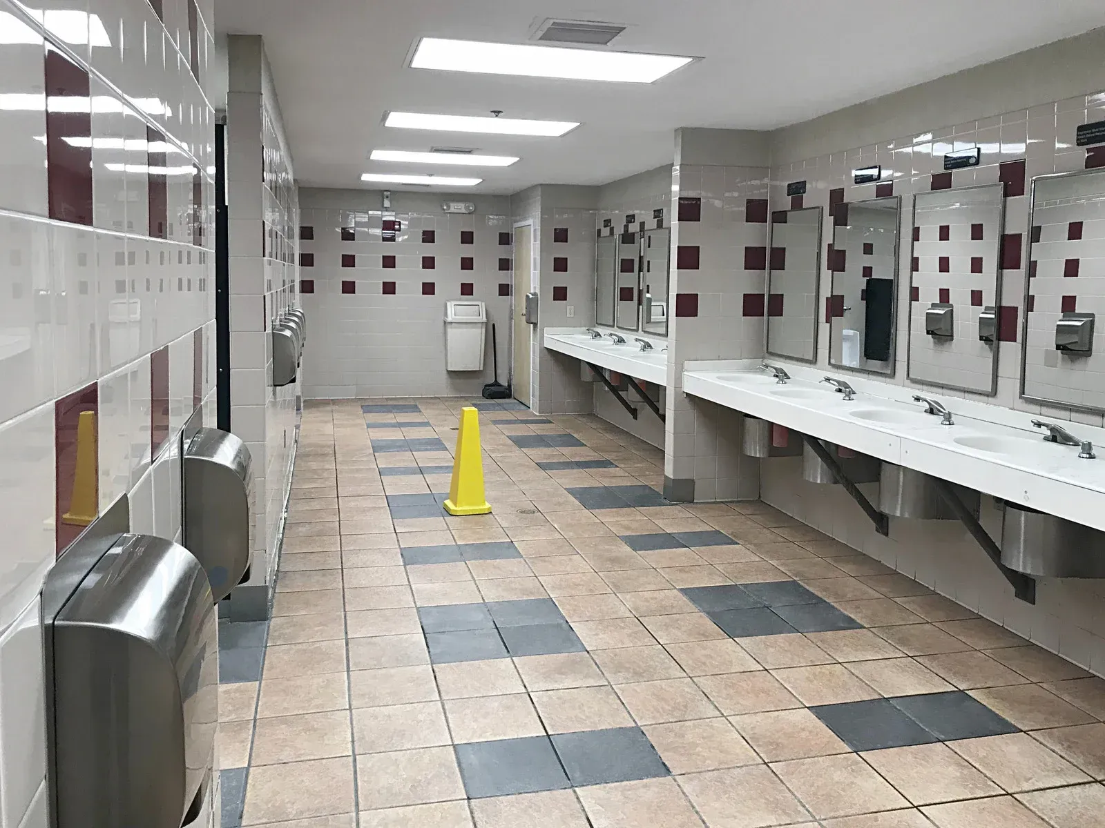 Public restroom with multiple sinks, mirrors, and hand dryers, with tiled walls and floor. A caution cone sits in the middle.