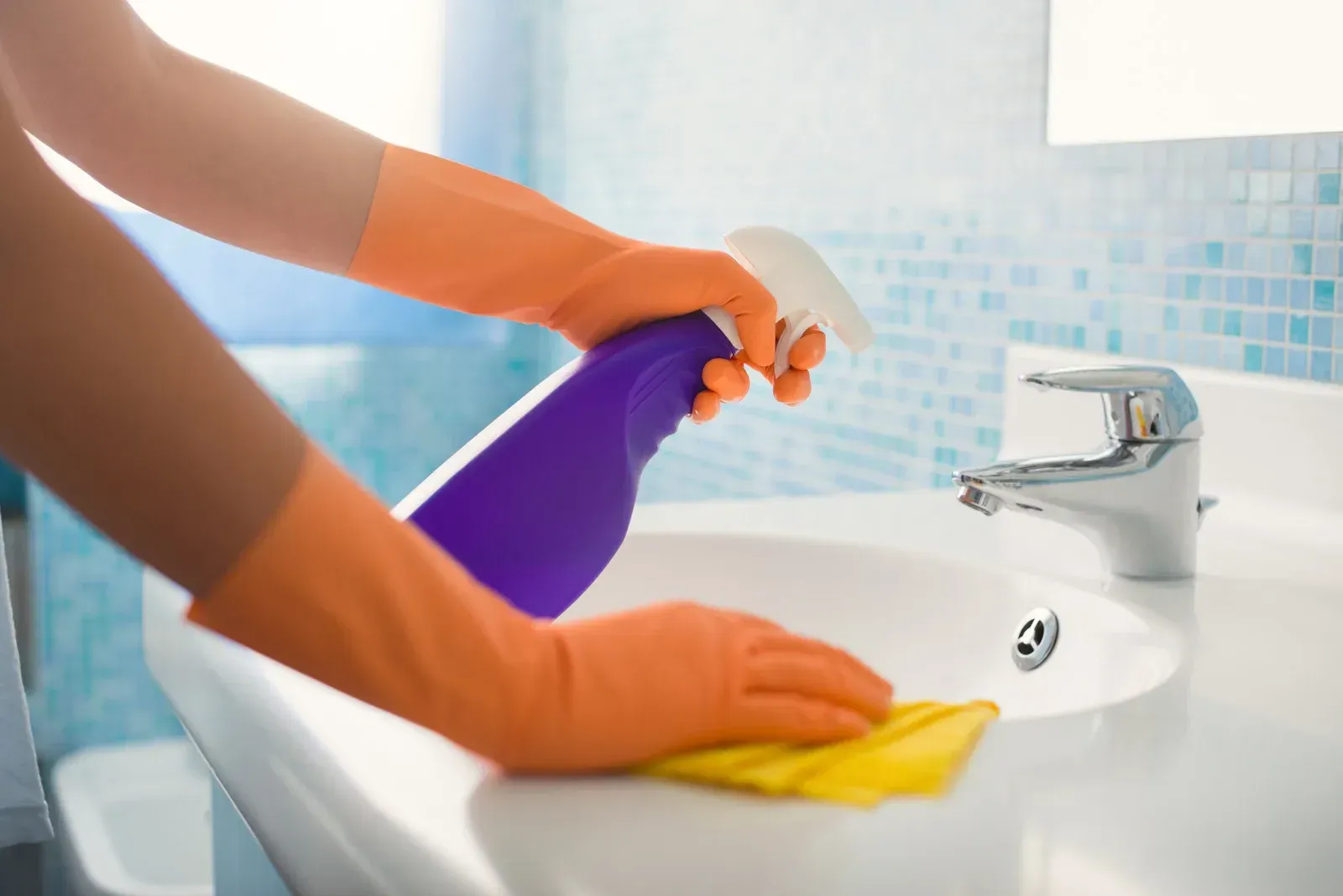 Hands in orange gloves cleaning a white bathroom sink with a spray bottle and yellow cloth.