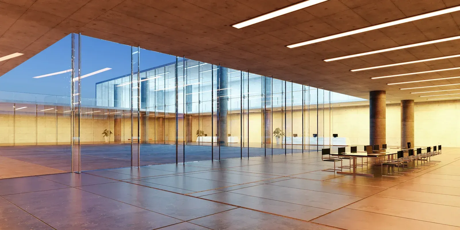 Modern interior with glass walls, concrete ceiling, and a long table with chairs. Sunlight streams through.