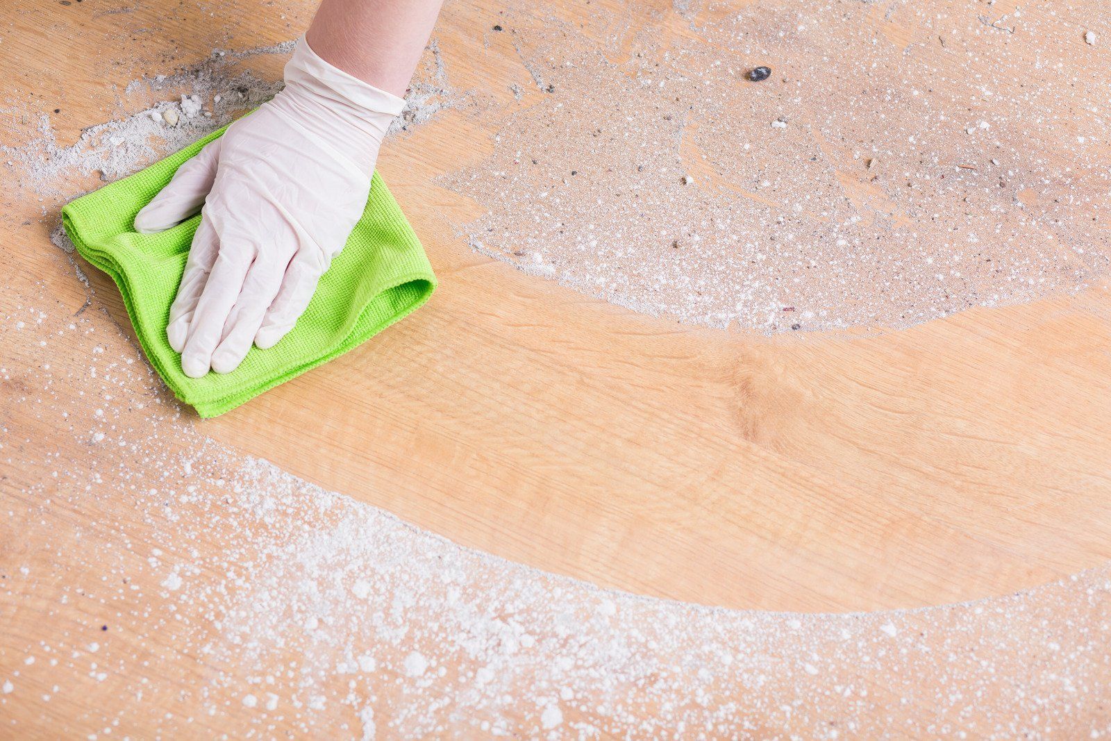 Gloved hand wiping white powder from a light wood surface with a green cloth.