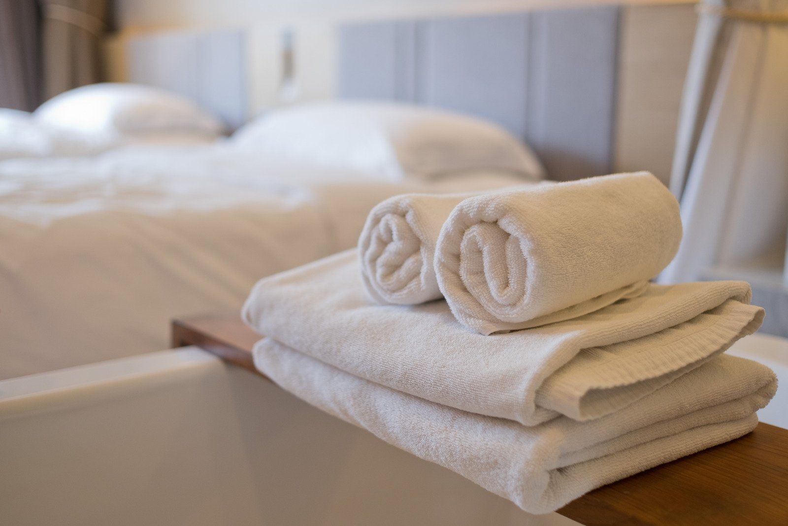 White rolled and folded towels on a wooden tray, next to a white bathtub with a bed in the background.