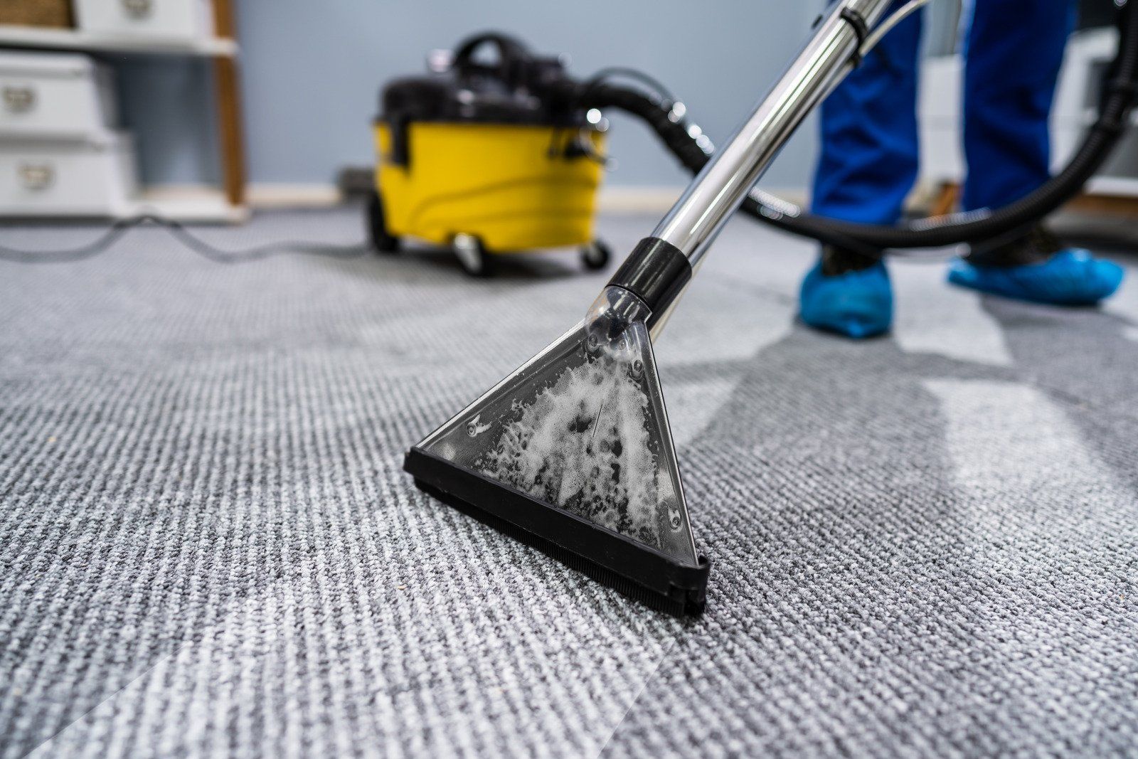 Person in blue pants vacuuming gray carpet with a yellow cleaning machine.
