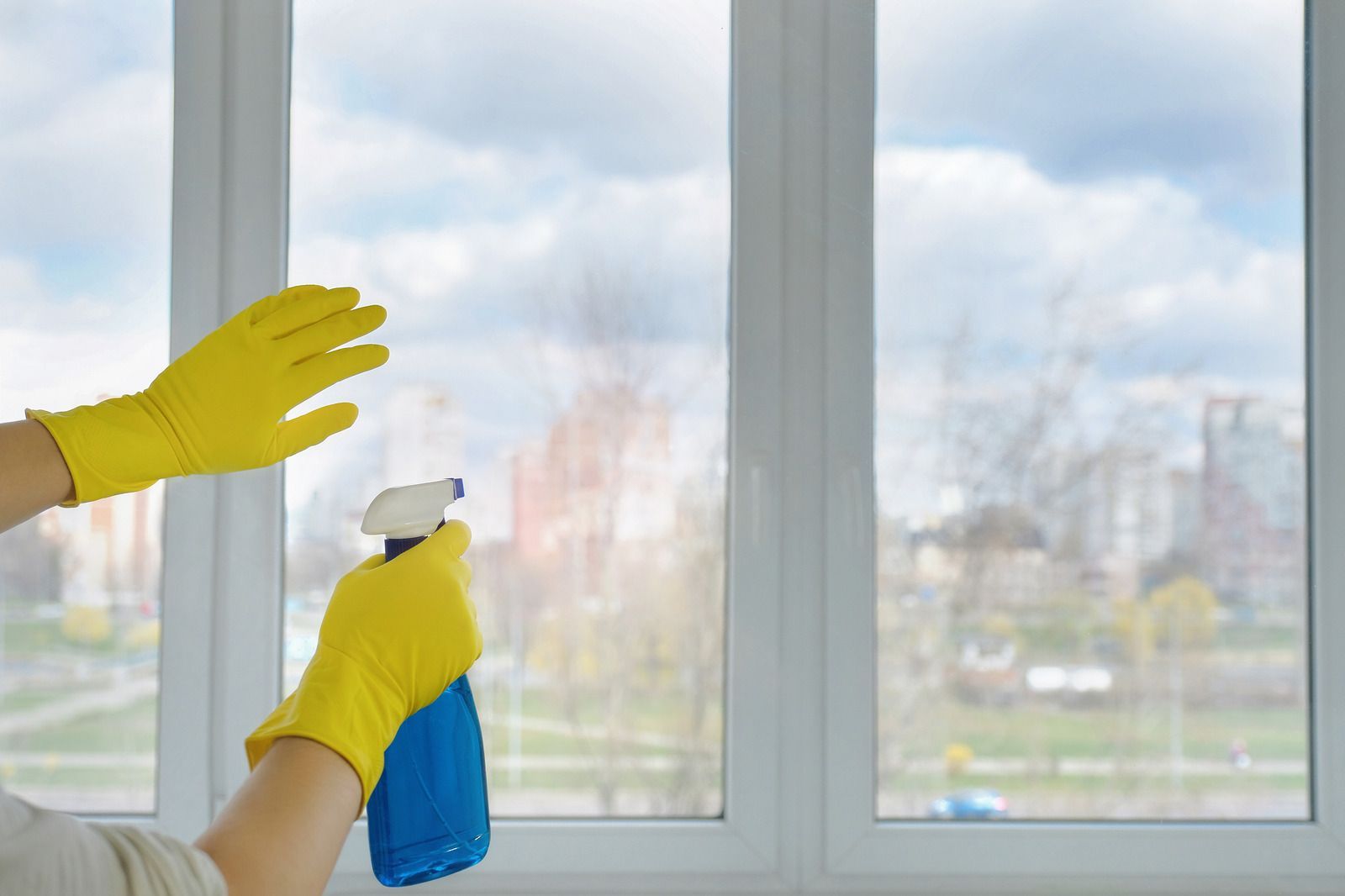 Person wearing yellow gloves spraying a window with blue bottle; city view in background.