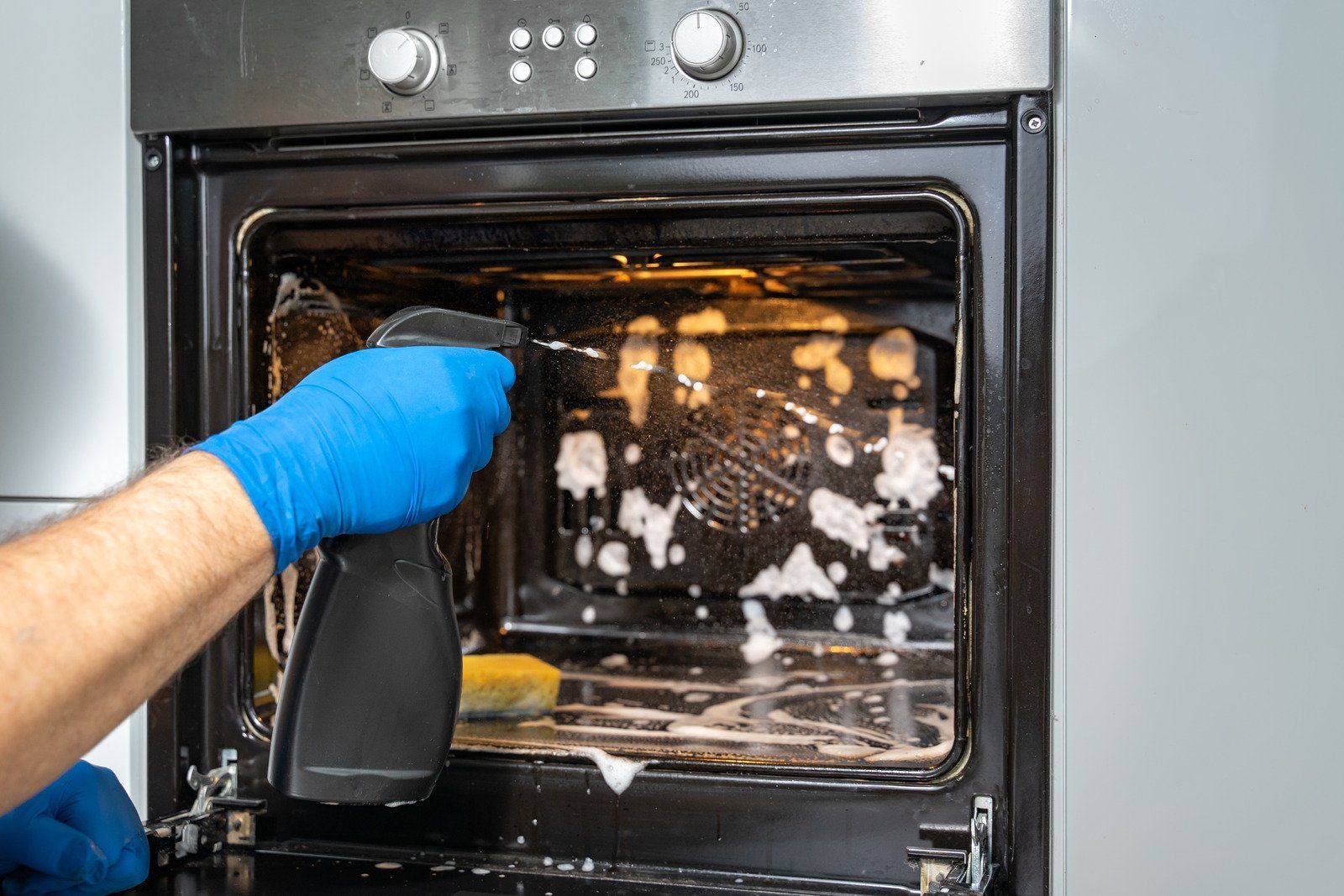 Person wearing blue gloves sprays cleaning solution into a dirty oven.