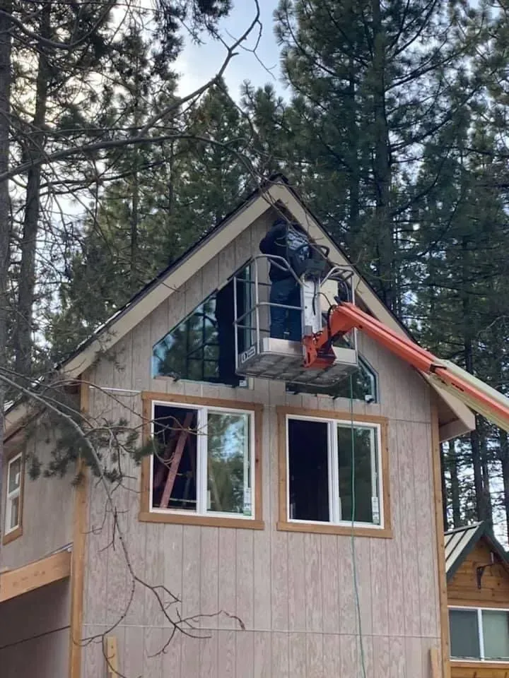 Worker in a lift painting the upper gable of a cabin among tall pine trees.