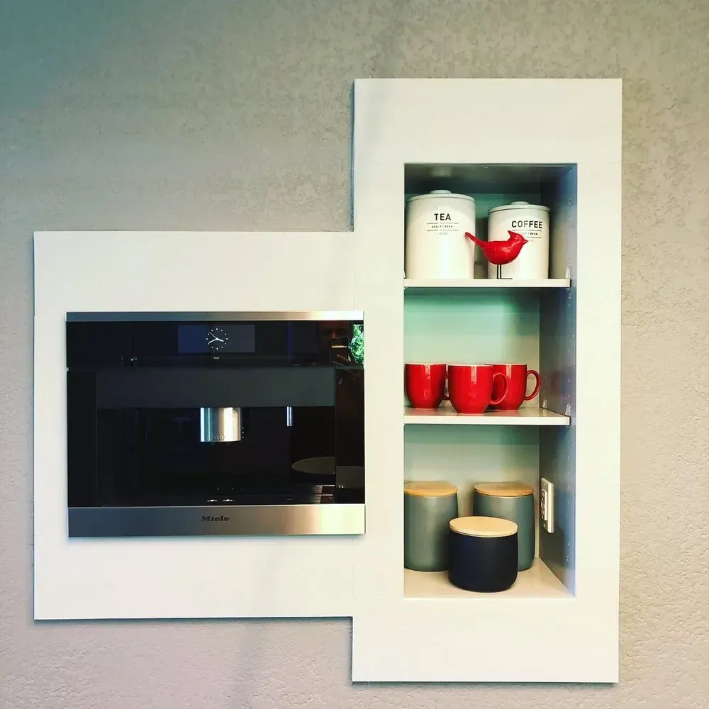 Built-in black oven beside white open shelving with mugs and cups on a light wall