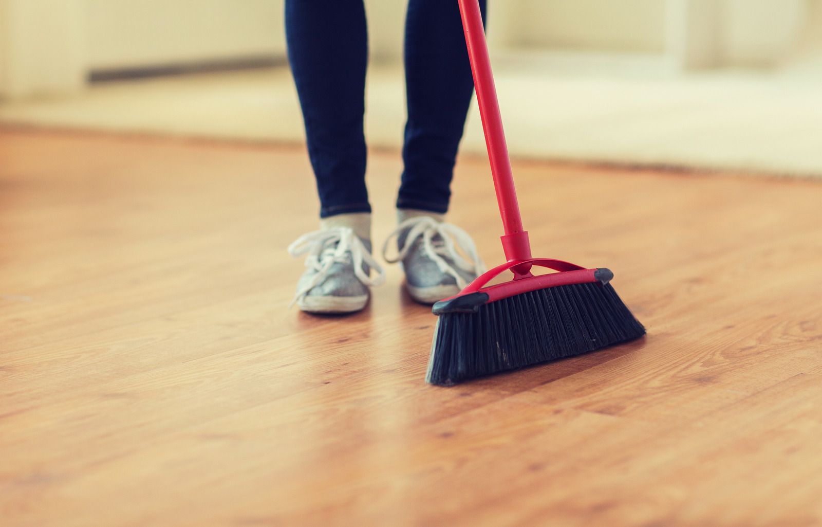 Person sweeping a wooden floor with a red broom, wearing gray sneakers and dark pants