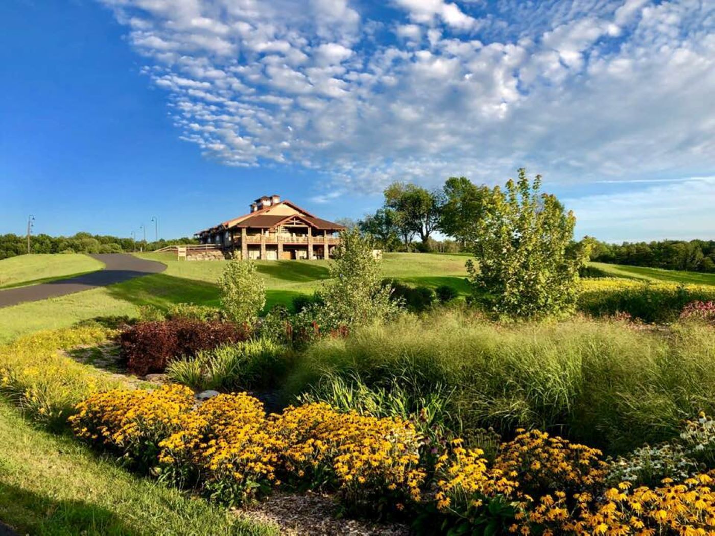 A large house is sitting on top of a grassy hill surrounded by flowers and trees.
