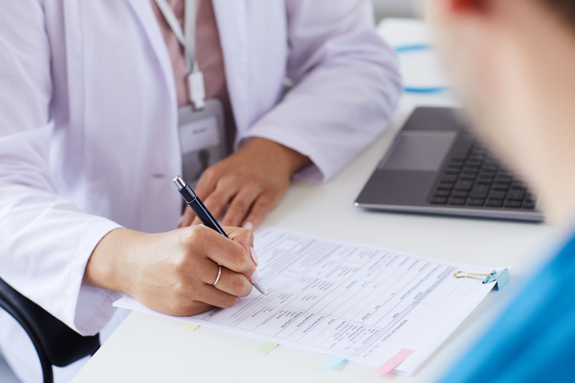 Doctor writing notes on a clipboard beside a laptop in a clinical office