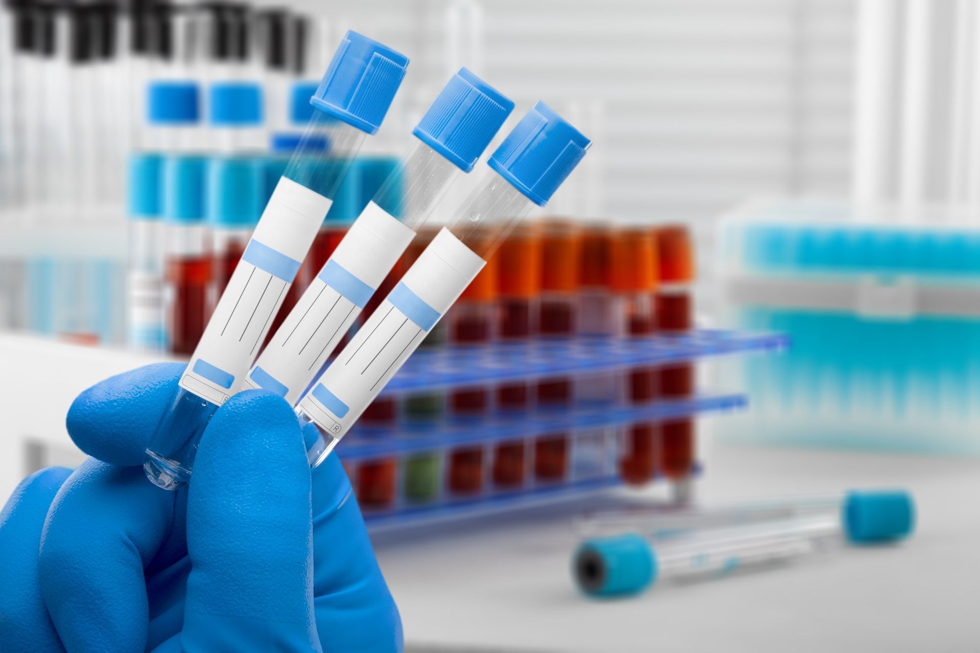 Gloved hand holding three blood sample tubes in a lab with racks of test tubes in the background Gloved hand holding three blood sample tubes in a lab with racks of test tubes in the background