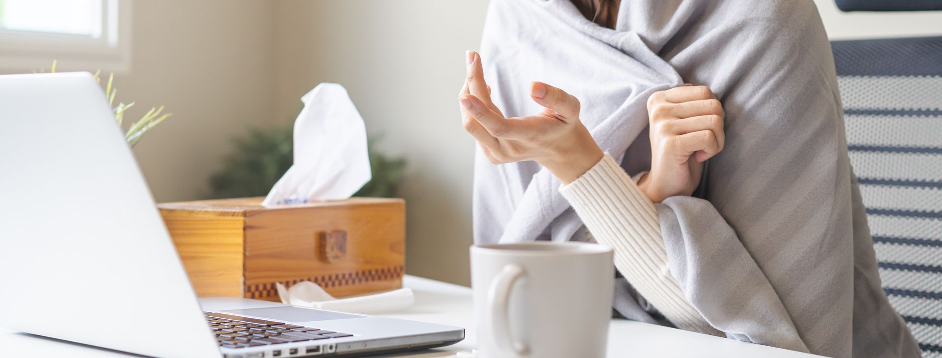 Person wrapped in a gray blanket at a desk, holding a tissue beside a laptop and mug. Person wrapped in a gray blanket at a desk, holding a tissue beside a laptop and mug.