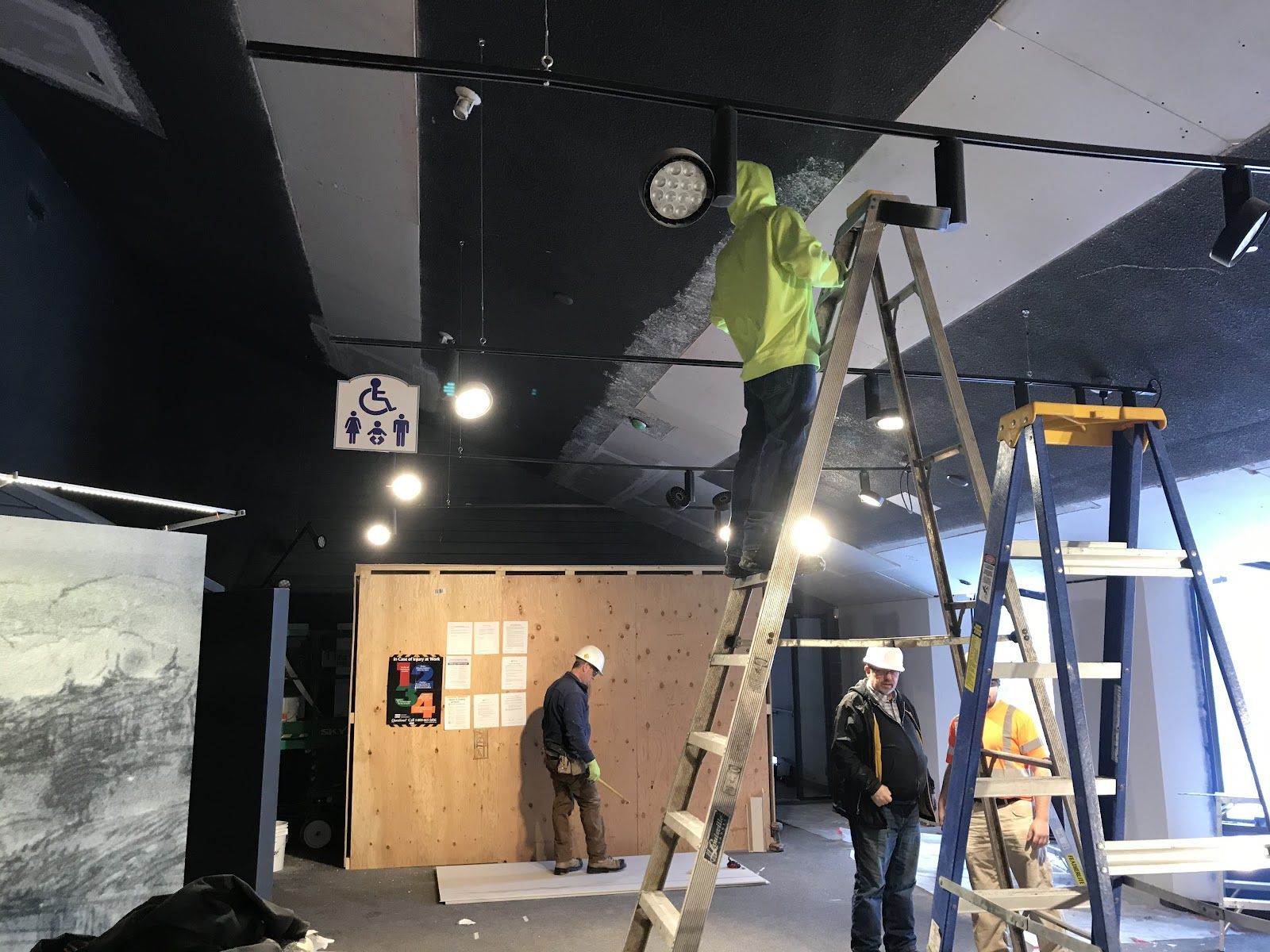 Workers installing ceiling fixtures in a room with ladders and tools under bright lights.
