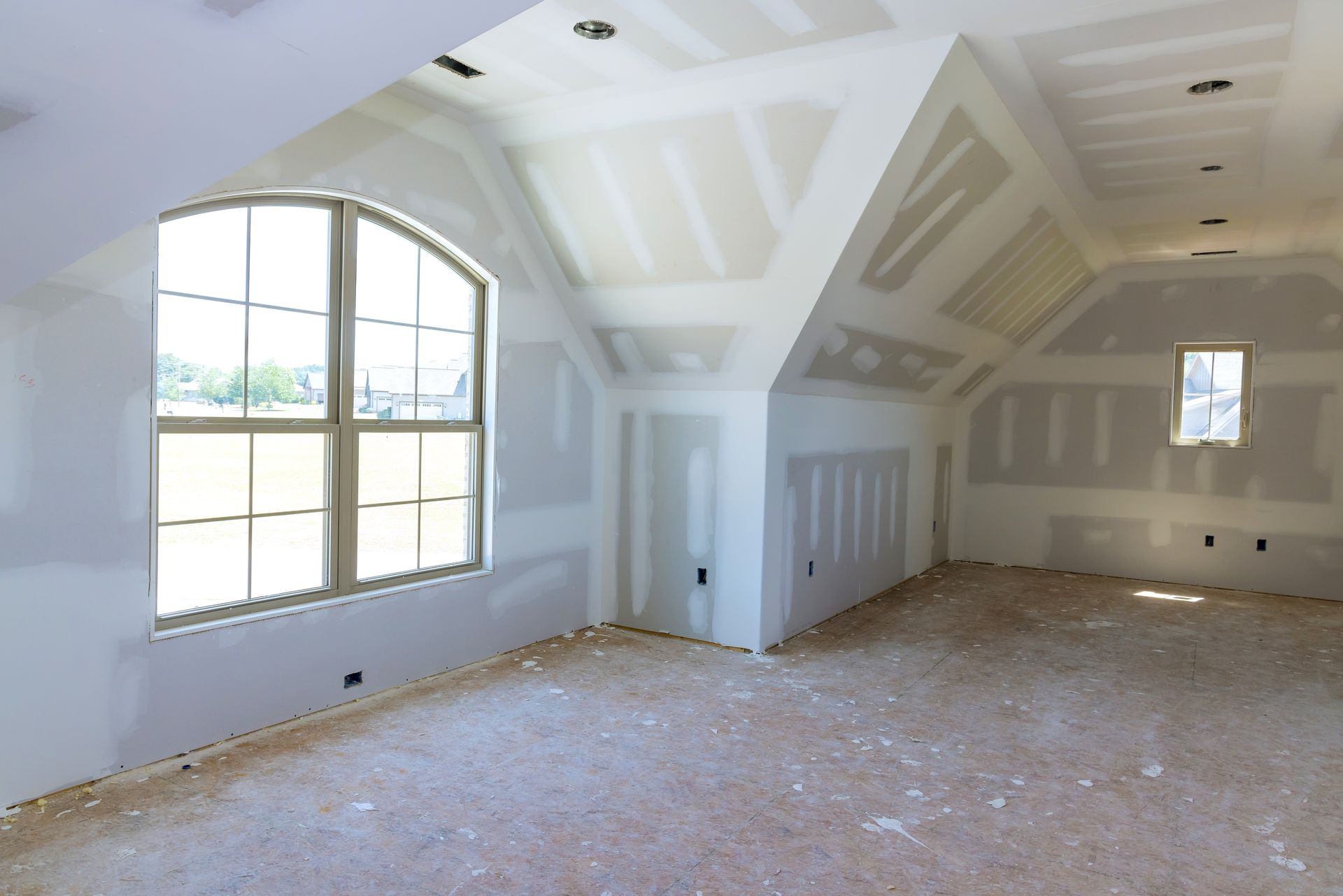 Empty unfinished room with vaulted white ceiling, large arched window, and exposed flooring debris.