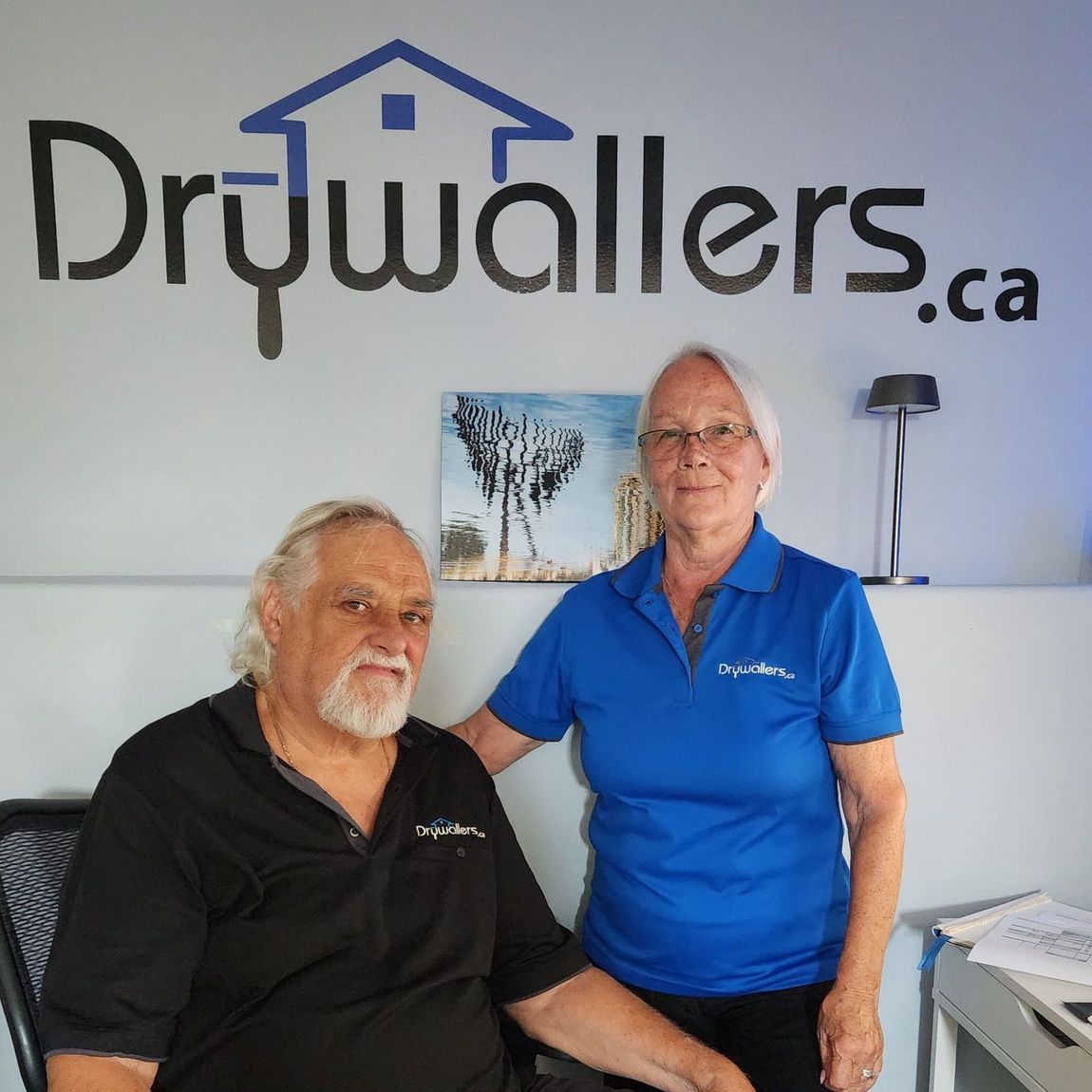 Two people in an office, in front of a drywall logo. Man seated, woman standing, both wearing branded shirts.