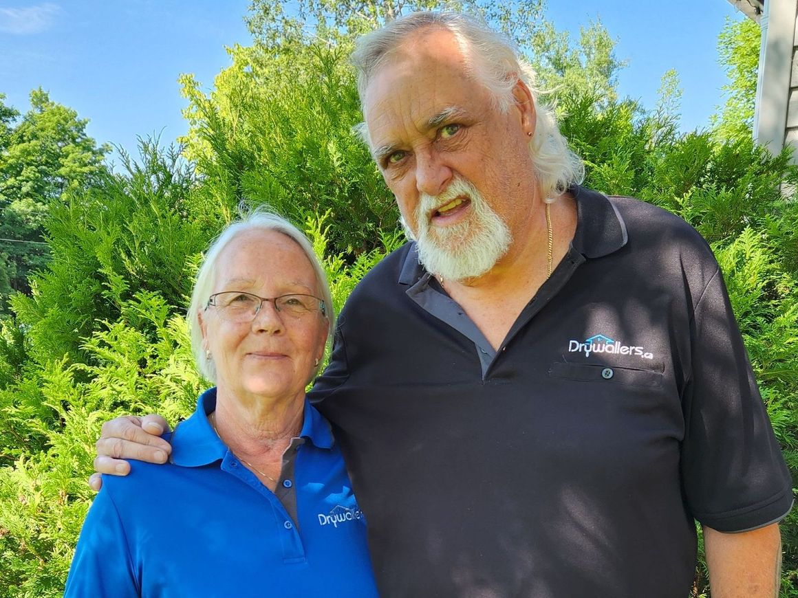 Two people stand outside; woman in blue shirt, man in black shirt with arm around woman.