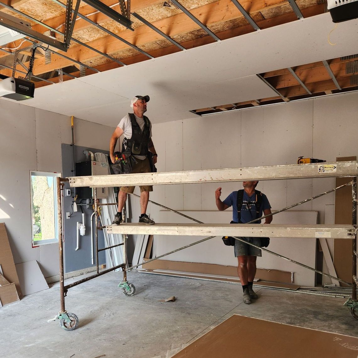 Two construction workers in a garage installing drywall, one on a scaffold, the other on the floor.