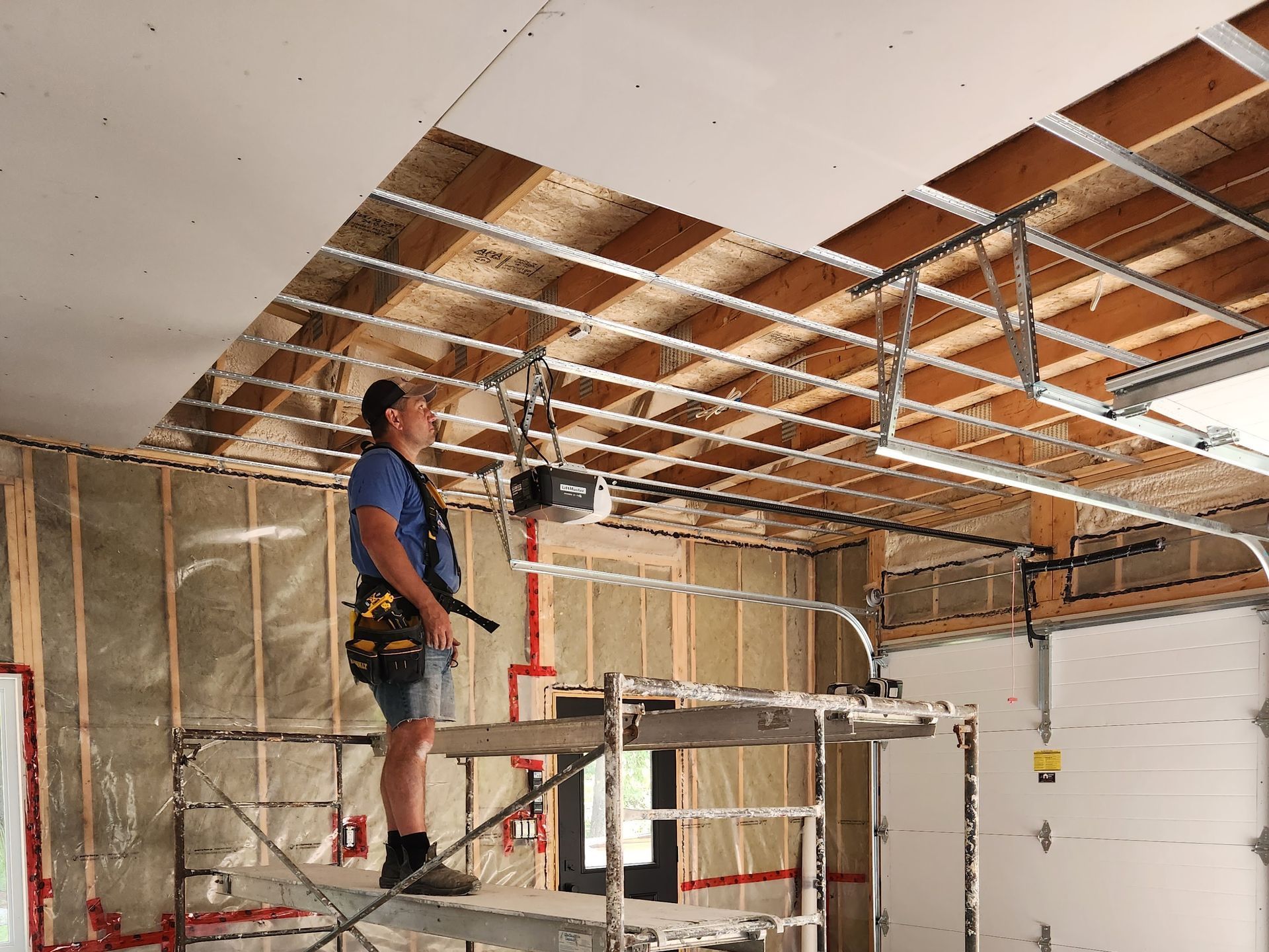 Man on scaffolding installing ceiling drywall in a garage under construction.