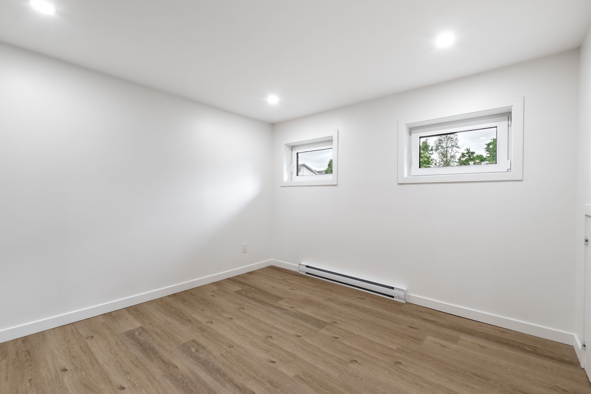 Empty white room with wood flooring, recessed lights, and two small windows.