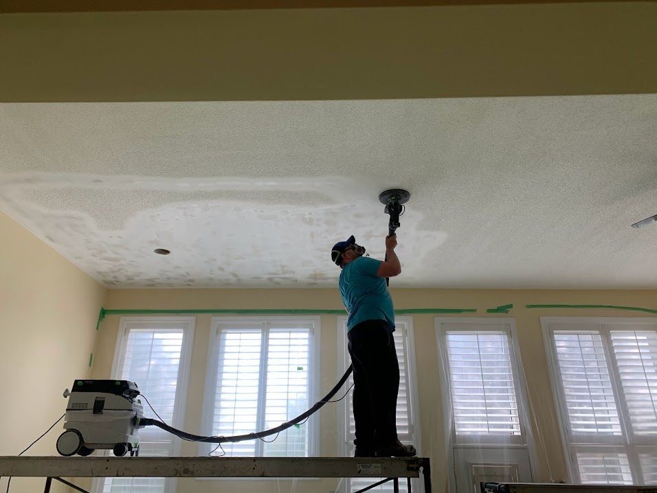 Man using a ceiling vacuum to clean a white textured ceiling in a room with windows and curtains