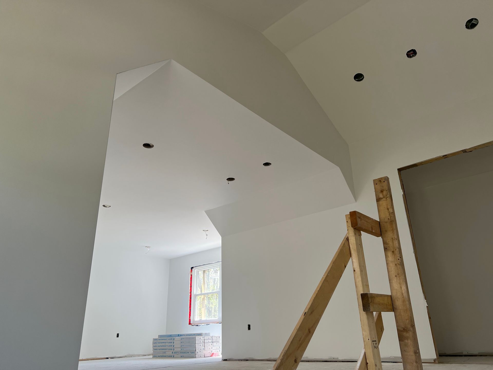 Interior view of a room under construction with white walls, angled ceiling, recessed lights, and a wooden ladder.
