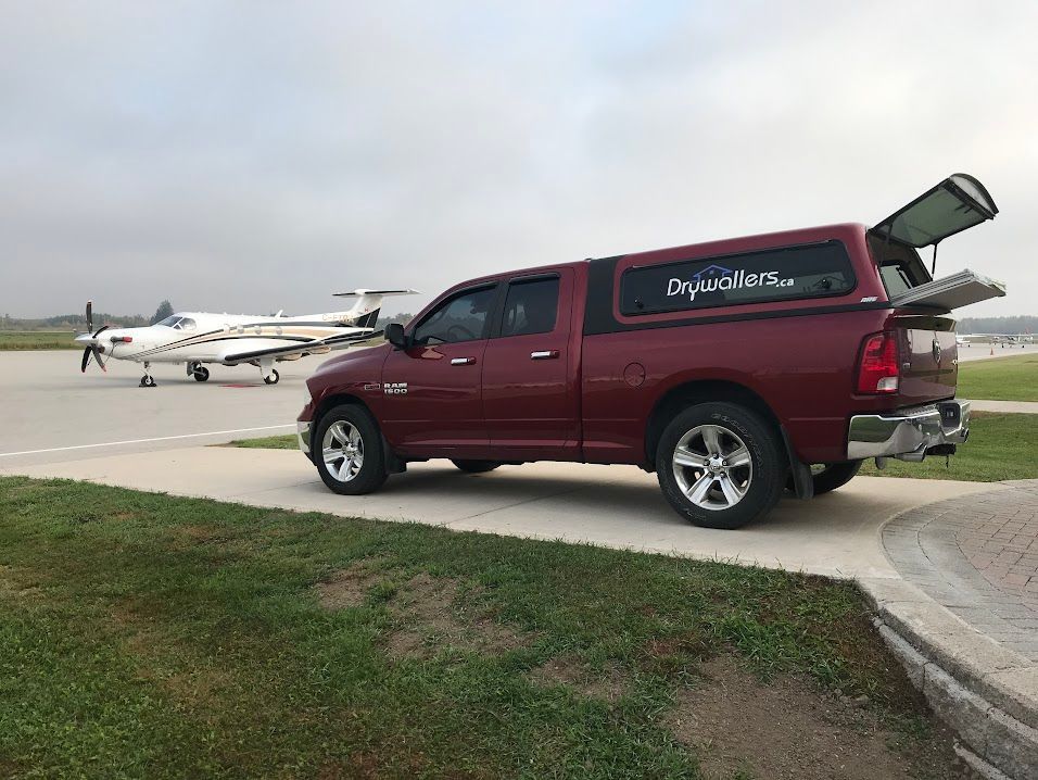 Red pickup truck with camper shell parked on a paved area near a small airplane on a runway.
