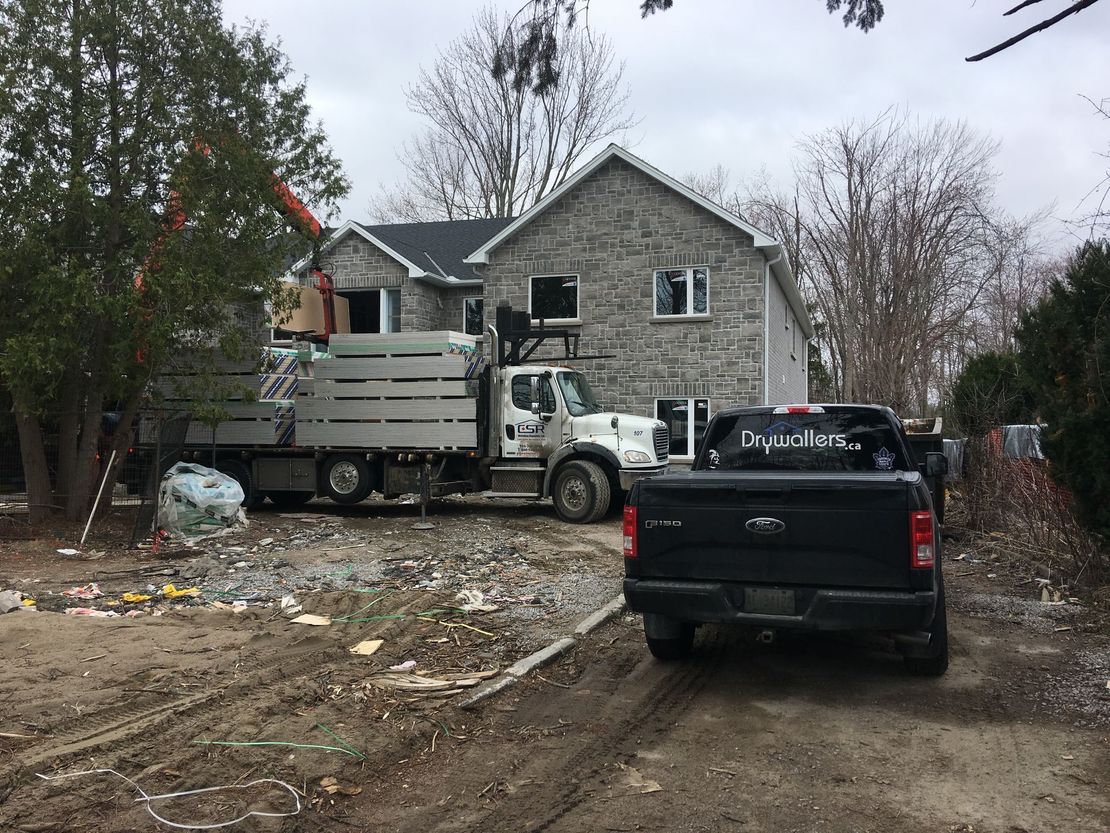 Trucks parked near a two-story house under construction. Materials are being unloaded; dirt ground. Cloudy sky.