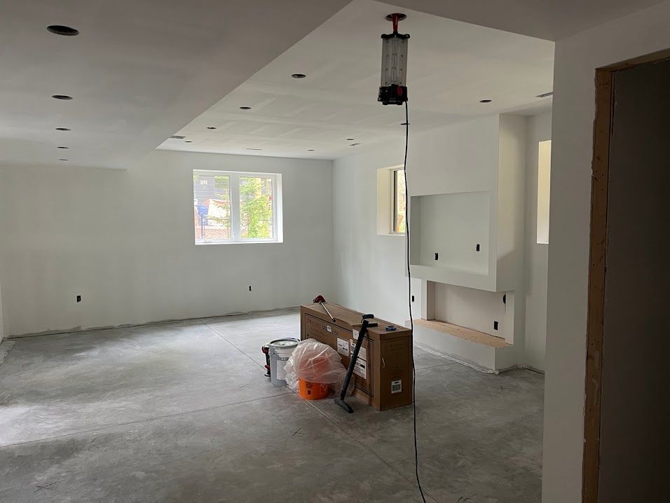 Interior of a room under construction, white walls, concrete floor, recessed lights, window, fireplace, and hanging work light.