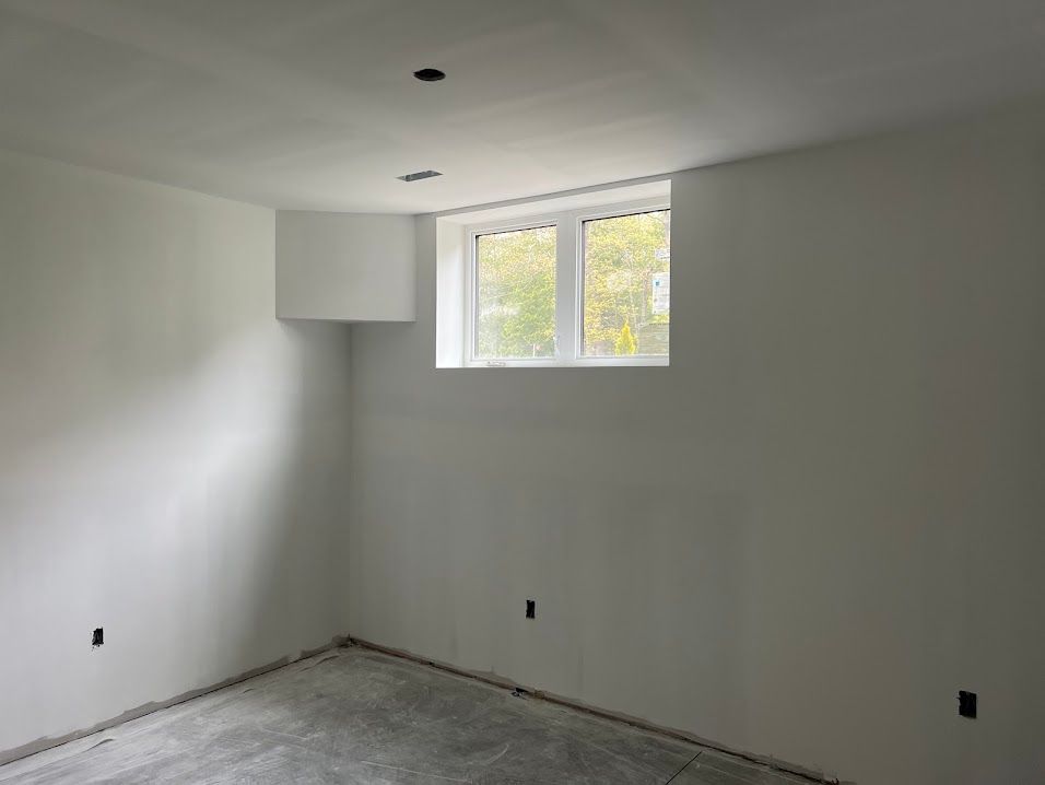 Empty white-walled room with a window, unfinished floor, and recessed ceiling lights.