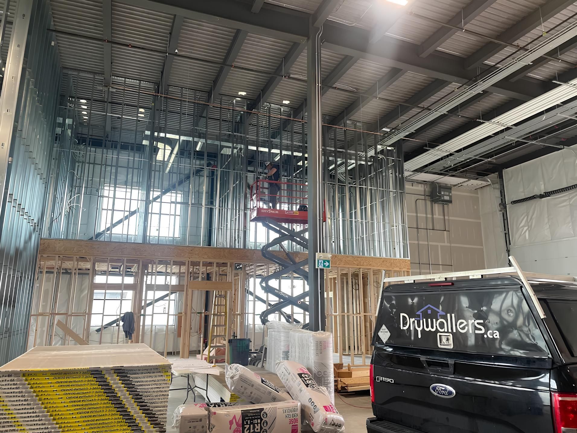 Interior construction site with metal framework, lift, and drywall materials. A drywaller van is parked nearby.