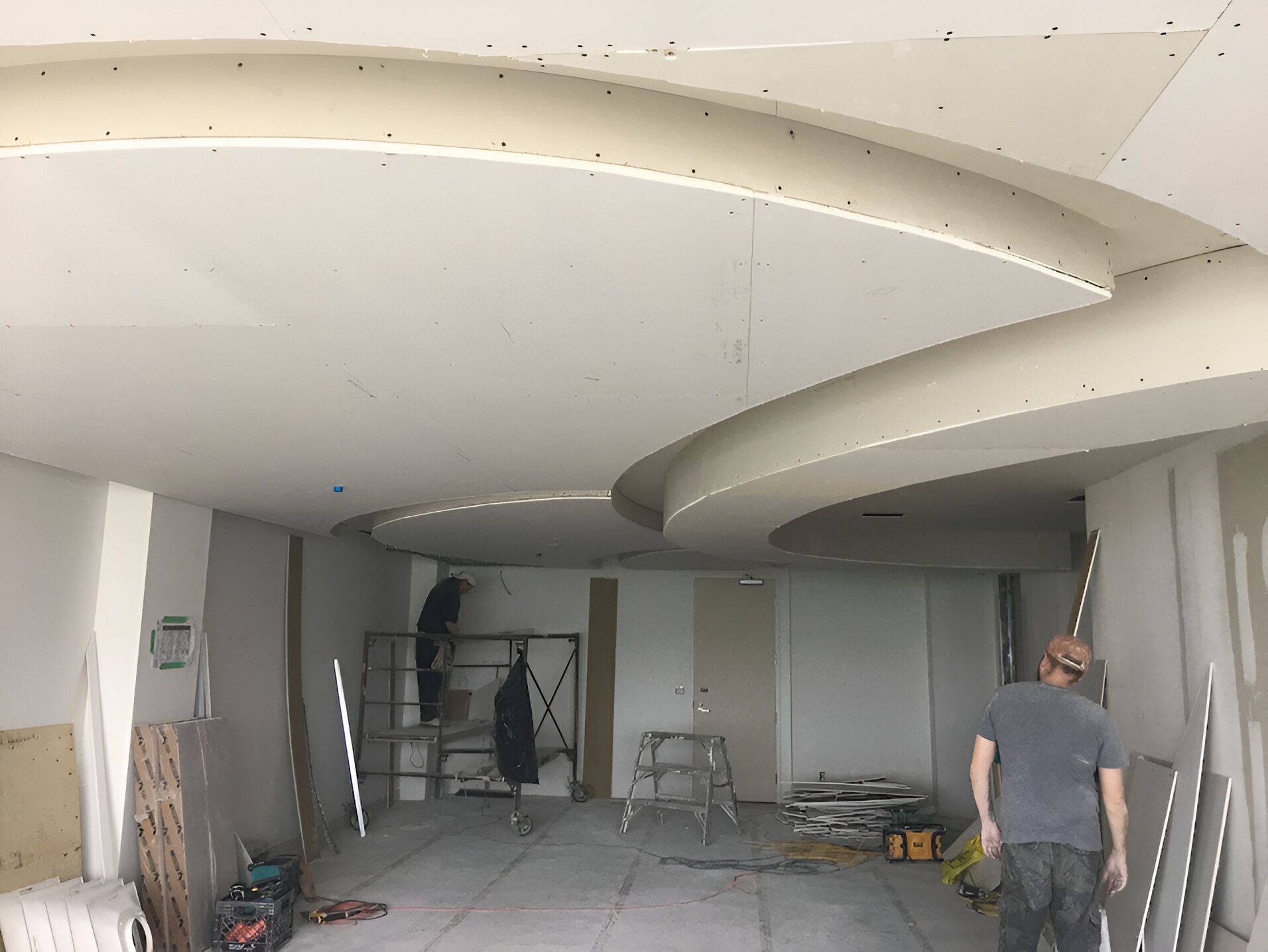 Workers installing drywall on a curved ceiling in a room under construction.