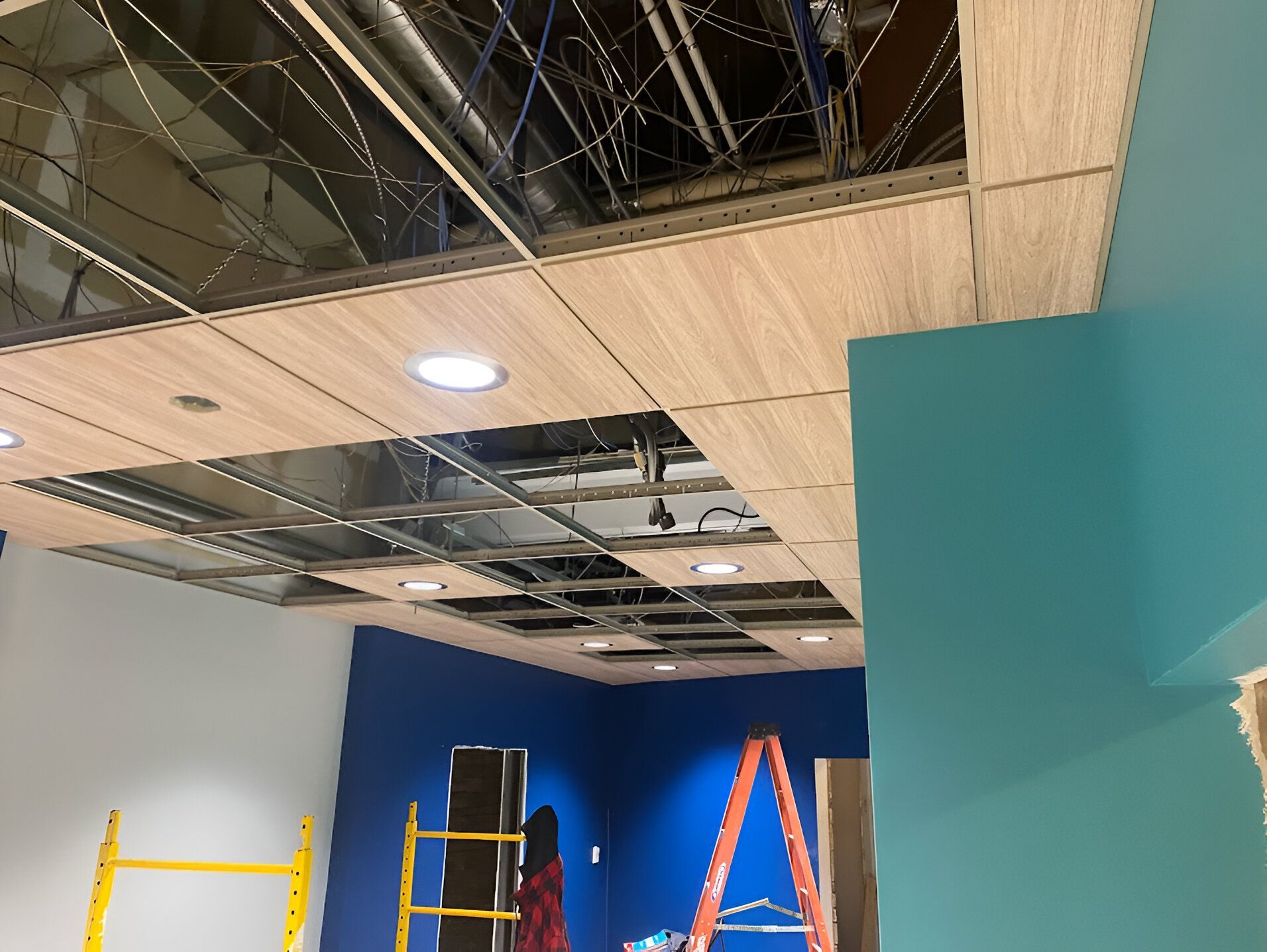 Ceiling under construction with exposed wiring, and partially installed wood-tone panels. Blue and white walls, with ladder visible.