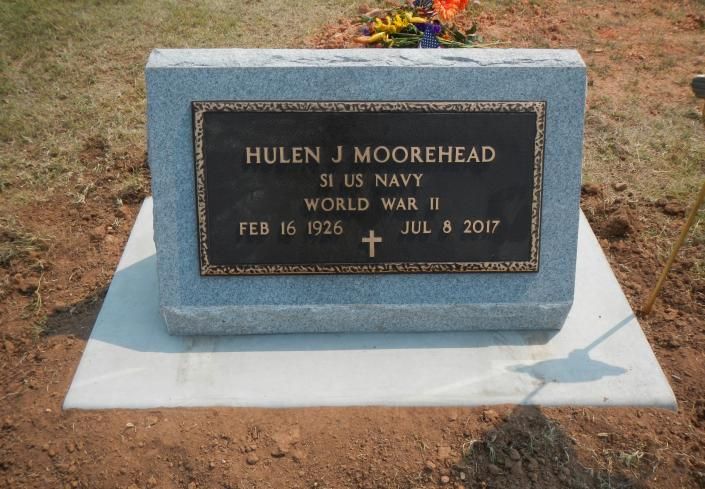 Blue gravestone with bronze plaque in a cemetery, surrounded by dry grass and flowers.