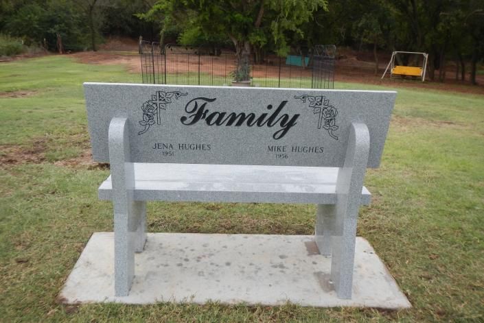 Gray memorial bench with “Family” engraved, set on a concrete pad in a grassy park.