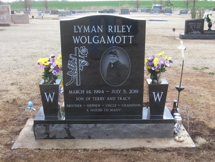Black granite gravestone with flowers at a cemetery, engraved with “LYMAN RILEY WOLGAMOTT” and a portrait.